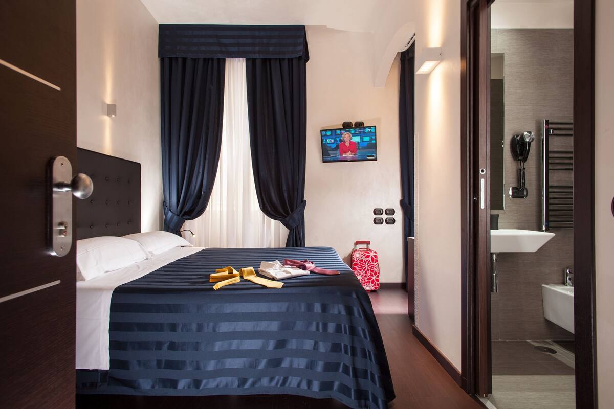 A hotel room with a bed, a television, a red suitcase, and a bathroom visible through an open door.