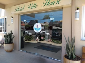 A entrada do Hotel Ville House, com porta de vidro, placa indicando 'Hotel Ville House Premium' e plantas em vasos na parte externa.