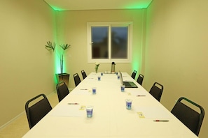 Uma sala de conferências com uma mesa comprida, cadeiras, um laptop e uma janela com persianas.