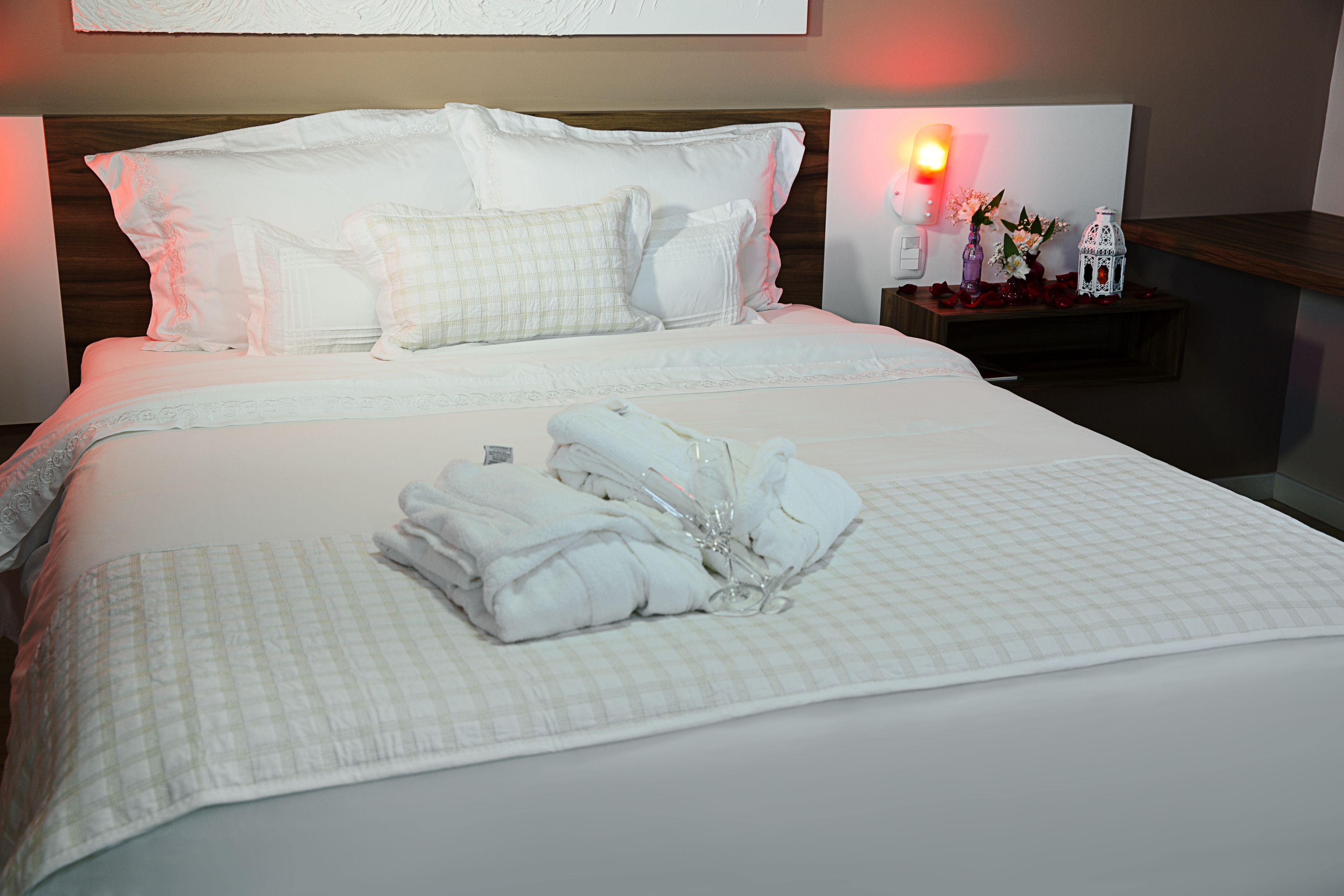 A neatly made bed with white linens and pillows, a bedside table with a vase and a lamp, and a wall-mounted light fixture.