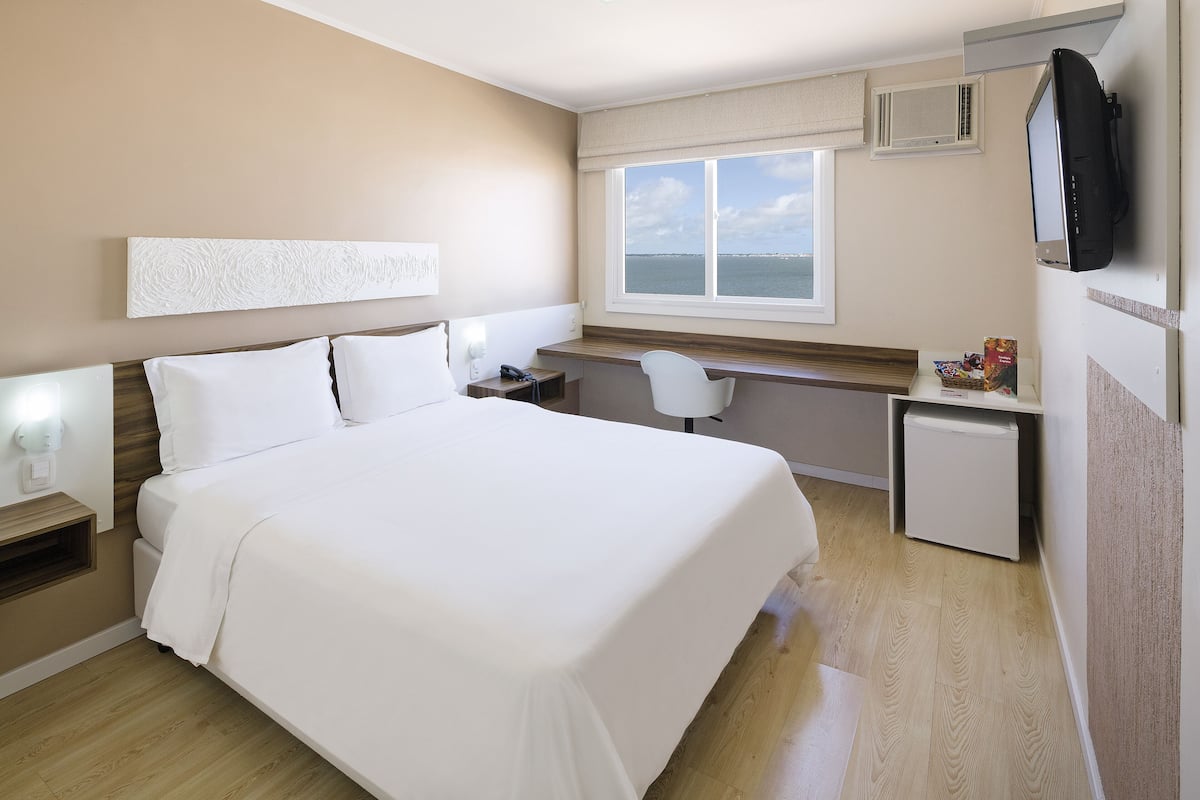 Ein Hotelzimmer mit einem großen Bett, einem Schreibtisch mit Stuhl, einem Fernseher und einem Fenster mit Meerblick.