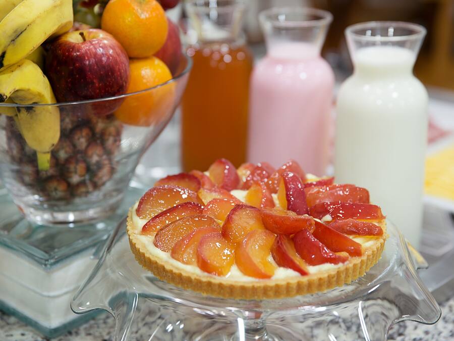 Uma tigela de frutas com bananas, maçãs e laranjas, um prato de vidro com uma torta de pêssego, e três garrafas de bebidas.