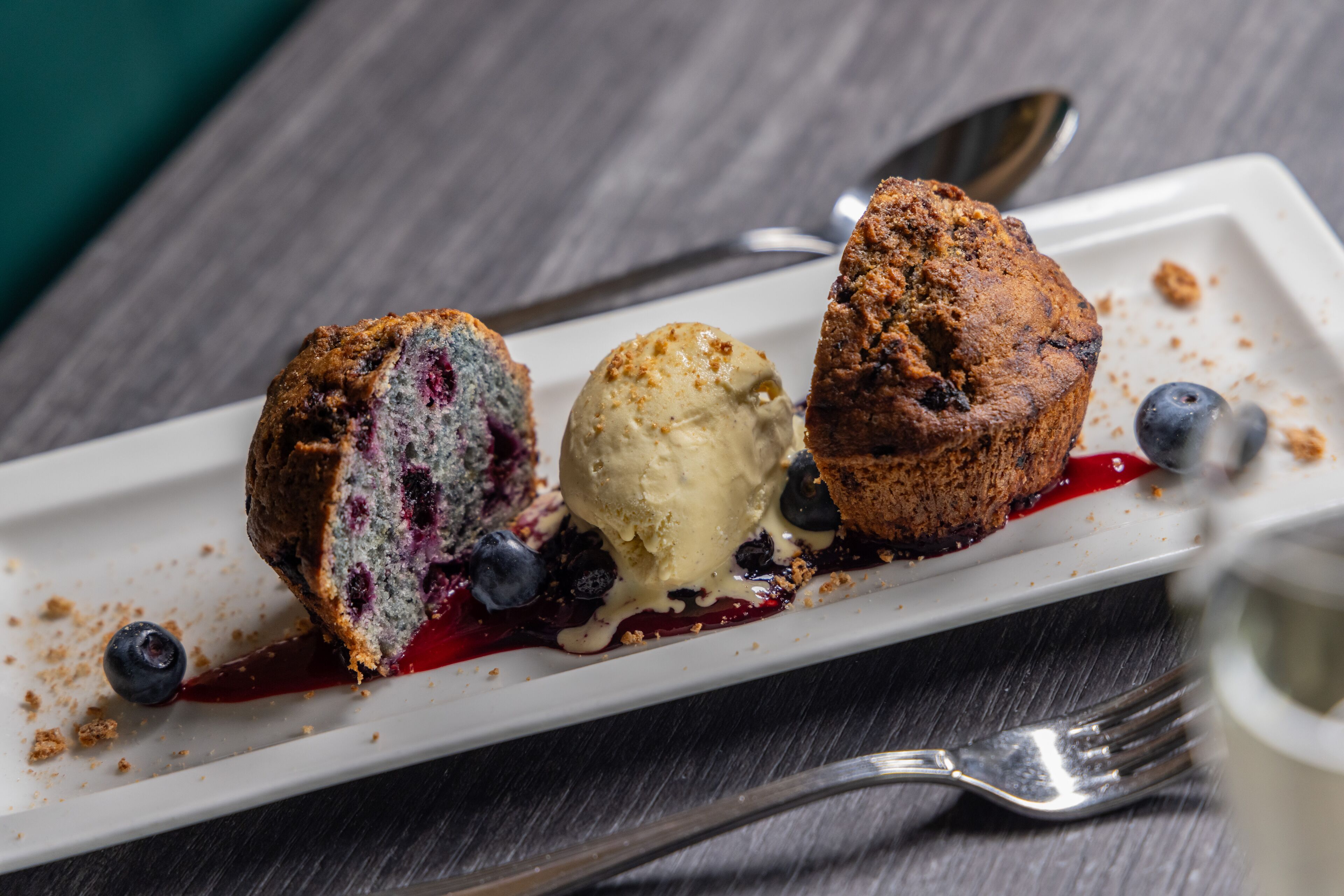 Un plato de postre con una porción de pastel de arándanos, una bola de helado de vainilla y salsa de arándanos.