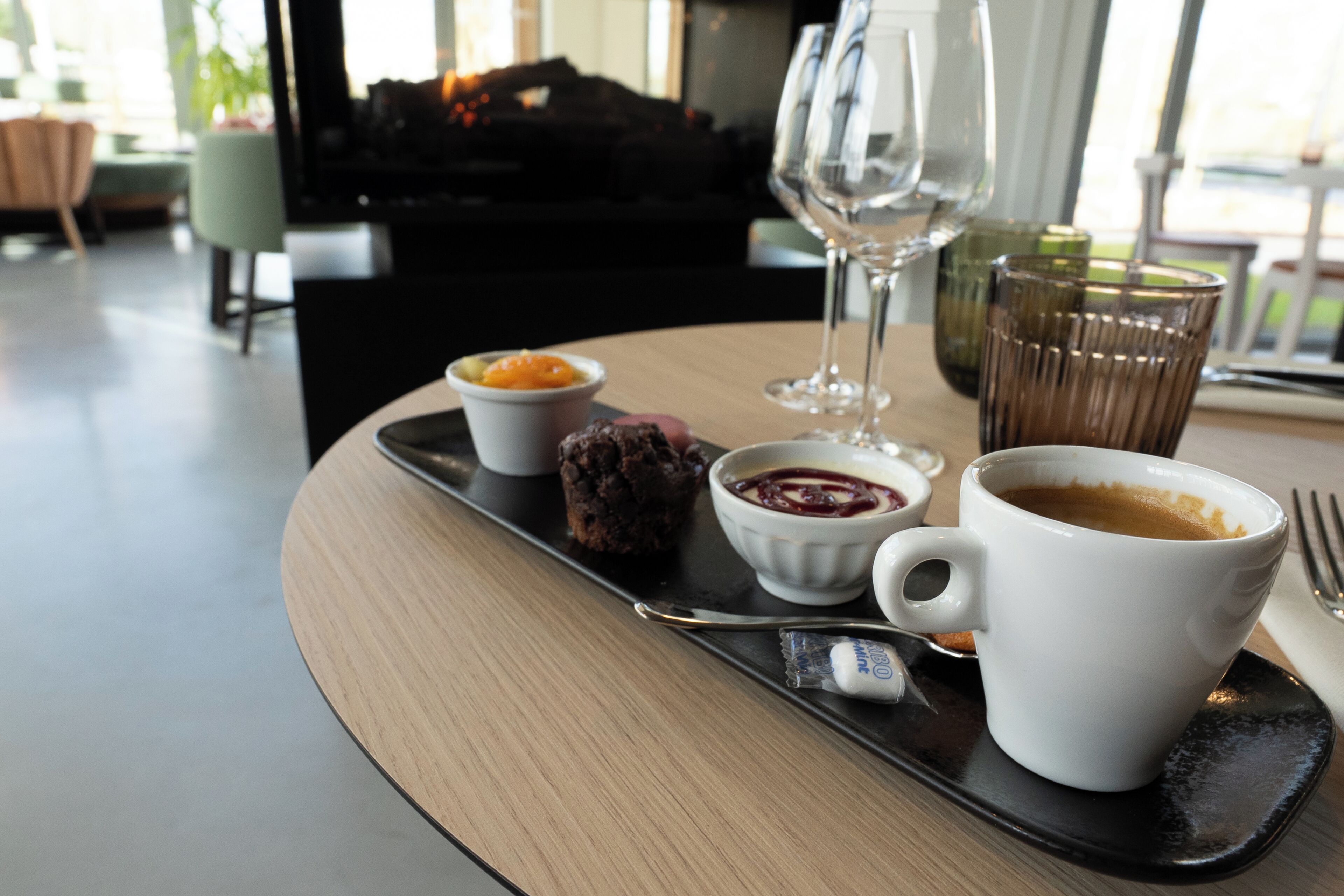 A tray with a cup of coffee, a chocolate dessert, and two small bowls of food, placed on a table in a room with a piano and a glass of wine in the background.