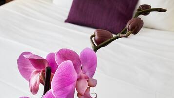 A hotel room with a neatly made bed, a purple pillow, and a pink orchid in the foreground.