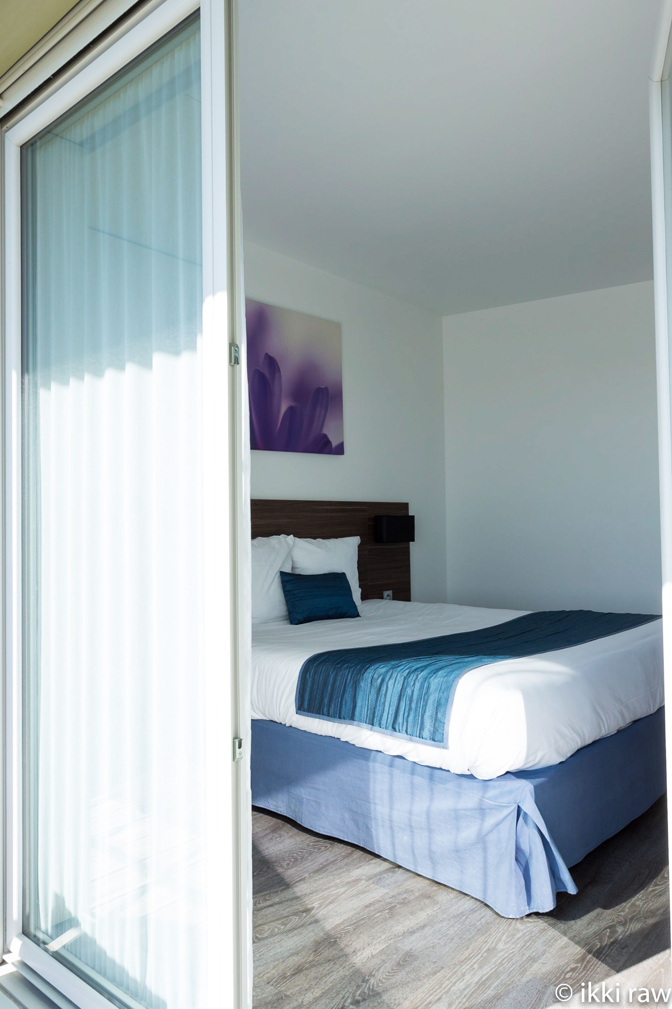 Une chambre d’hôtel avec un lit, une tête de lit, une œuvre d’art accrochée au mur et une fenêtre avec des stores.
