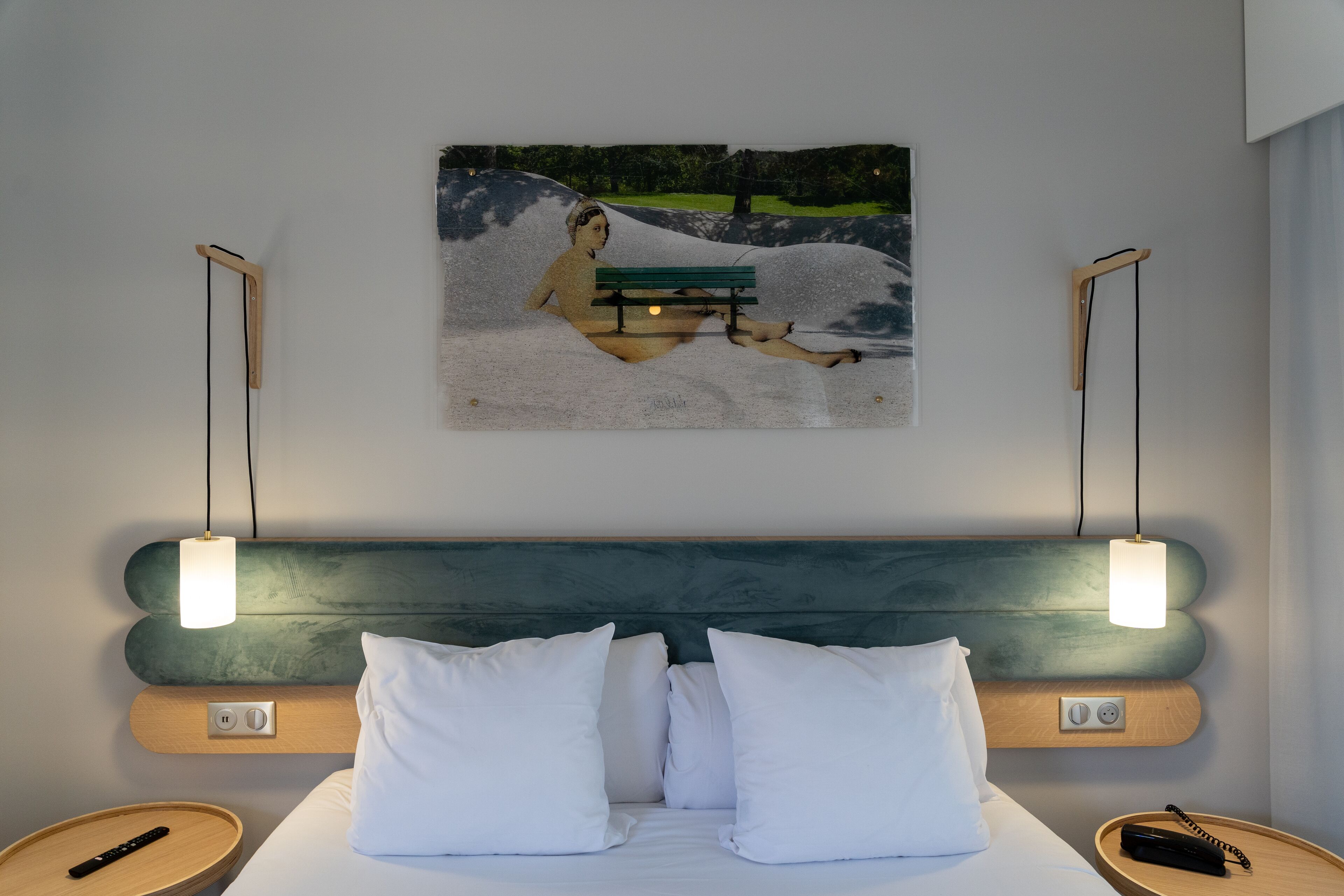 Habitación de hotel con cama, almohadas blancas, cabecero, mesitas de noche, control remoto y un cuadro de una escena de parque.