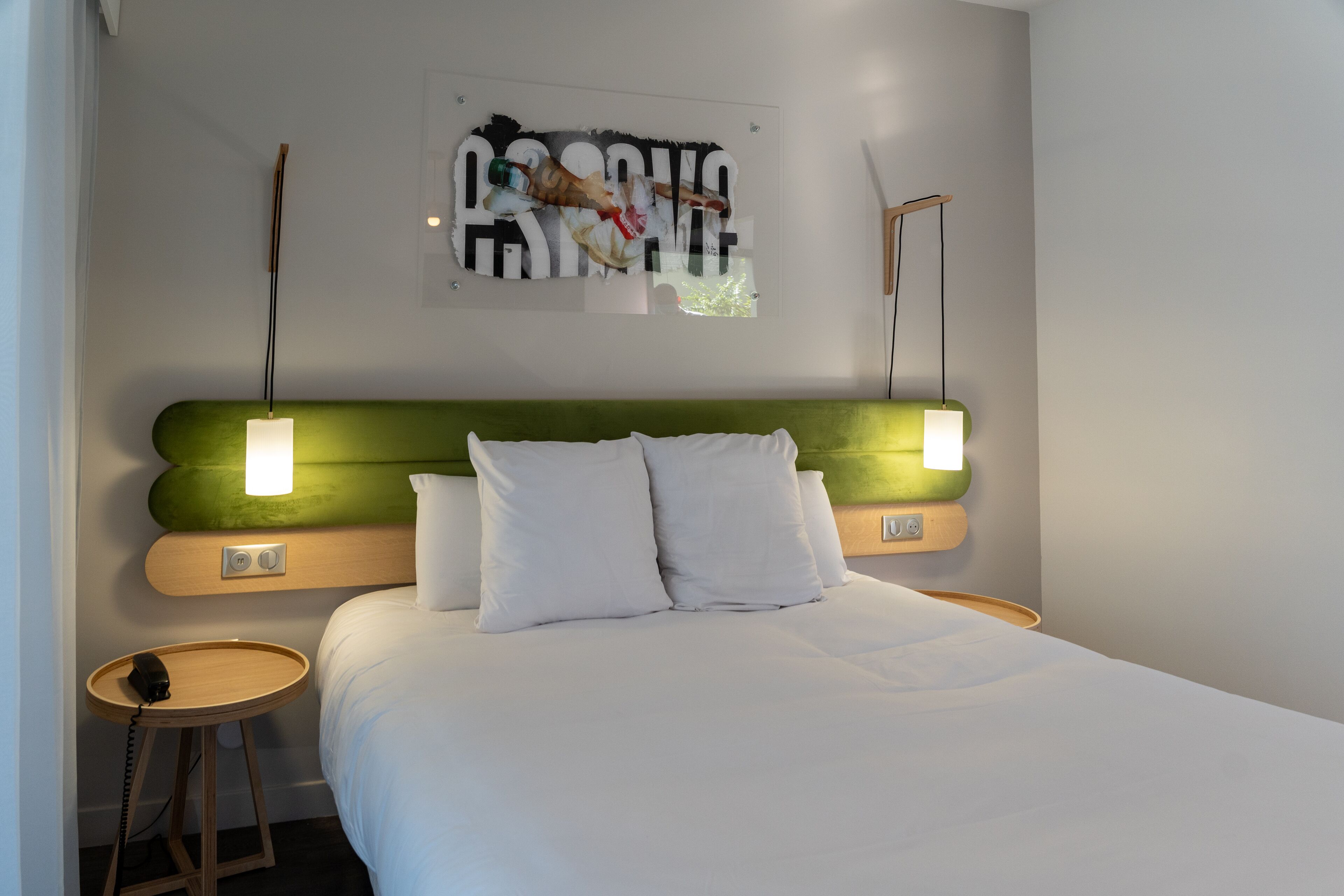 A modern hotel room with a green headboard, white bedding, a bedside table with a phone, and a wall-mounted artwork.