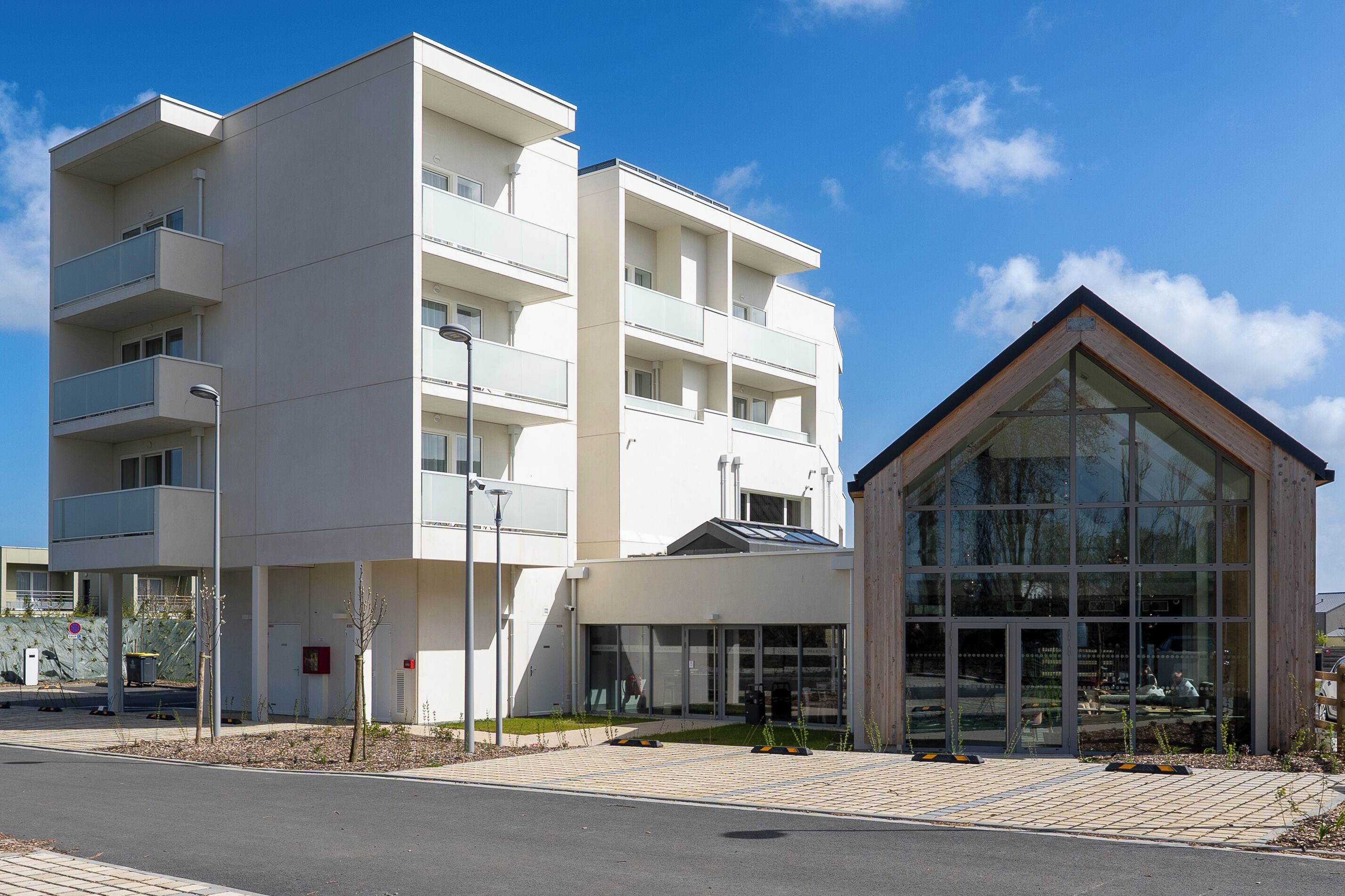 A modern multi-story building with balconies, a glass entrance, and a wooden structure to the right.