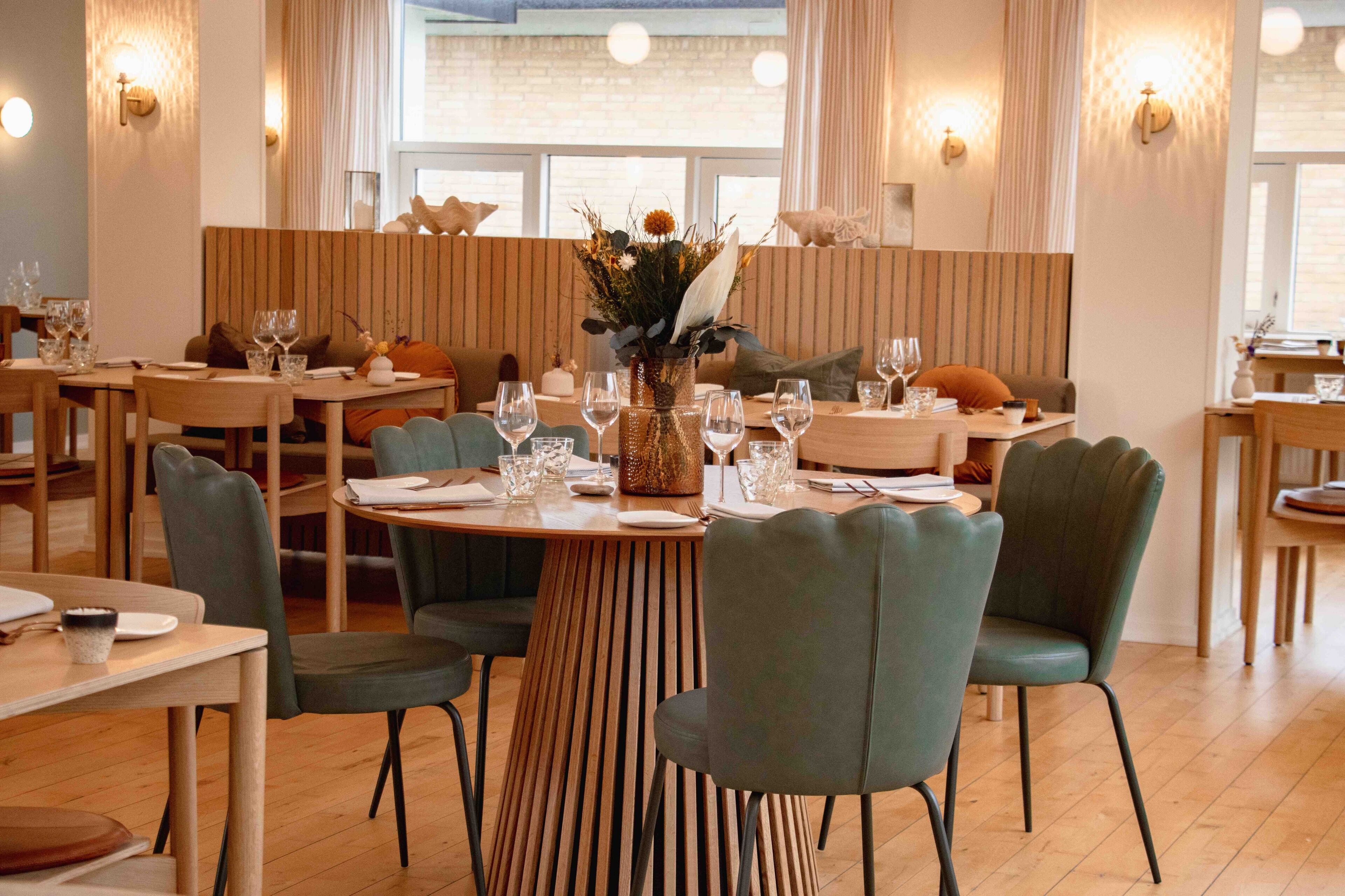 A restaurant with wooden tables, green upholstered chairs, and a central floral arrangement.