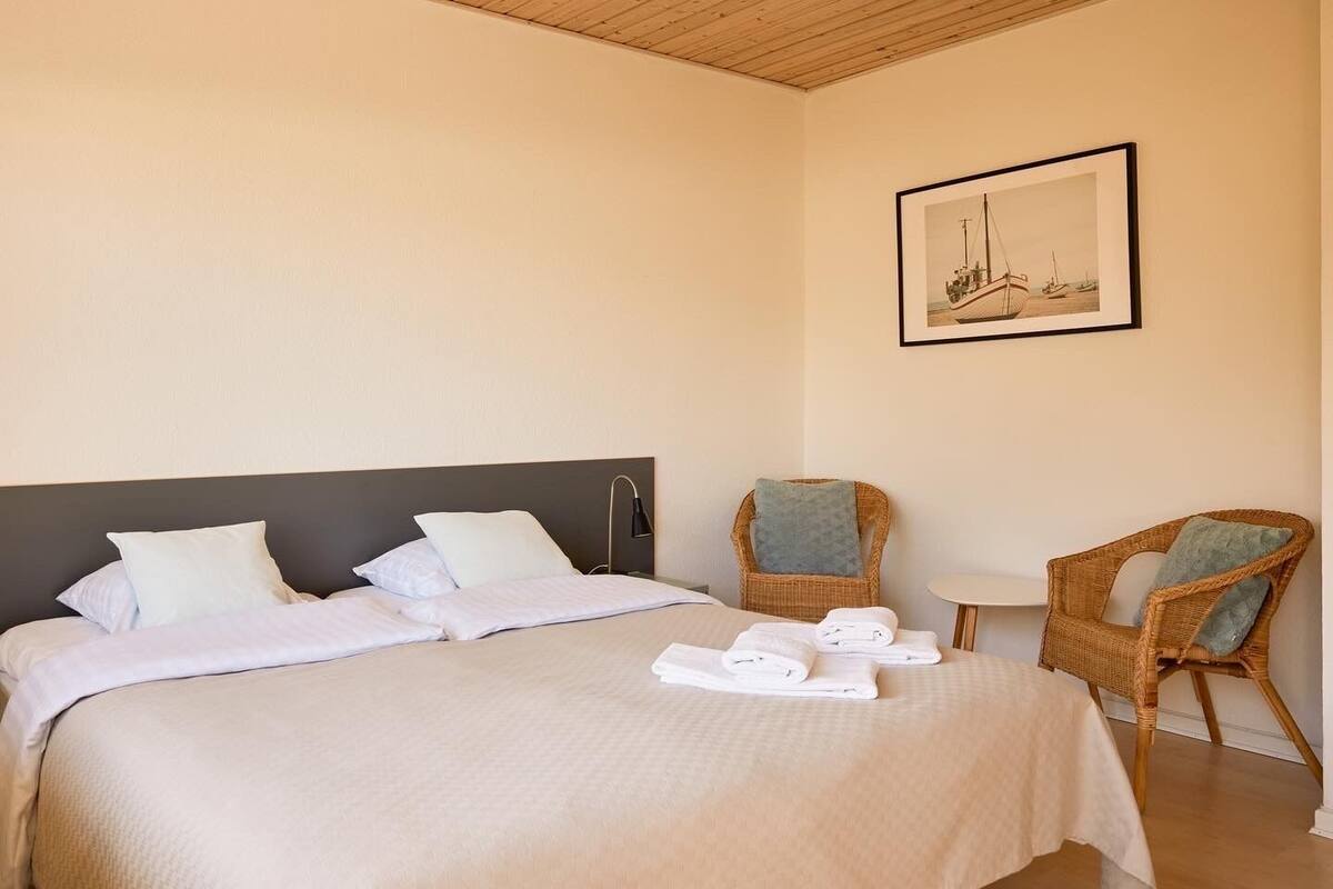 Una habitación de hotel con una cama, dos sillas de mimbre, una mesita y un cuadro en la pared.
