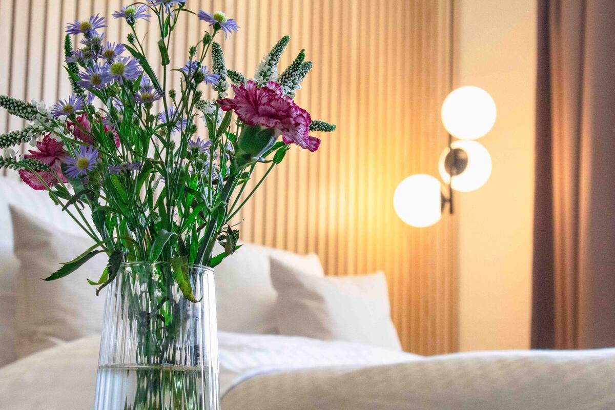 Vista clara de una habitación con una cama, una mesita de noche con un florero y una lámpara fijada a la pared.