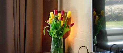 A hotel room with a wooden desk, a chair, a vase with tulips, a coffee maker, and a large window with curtains.
