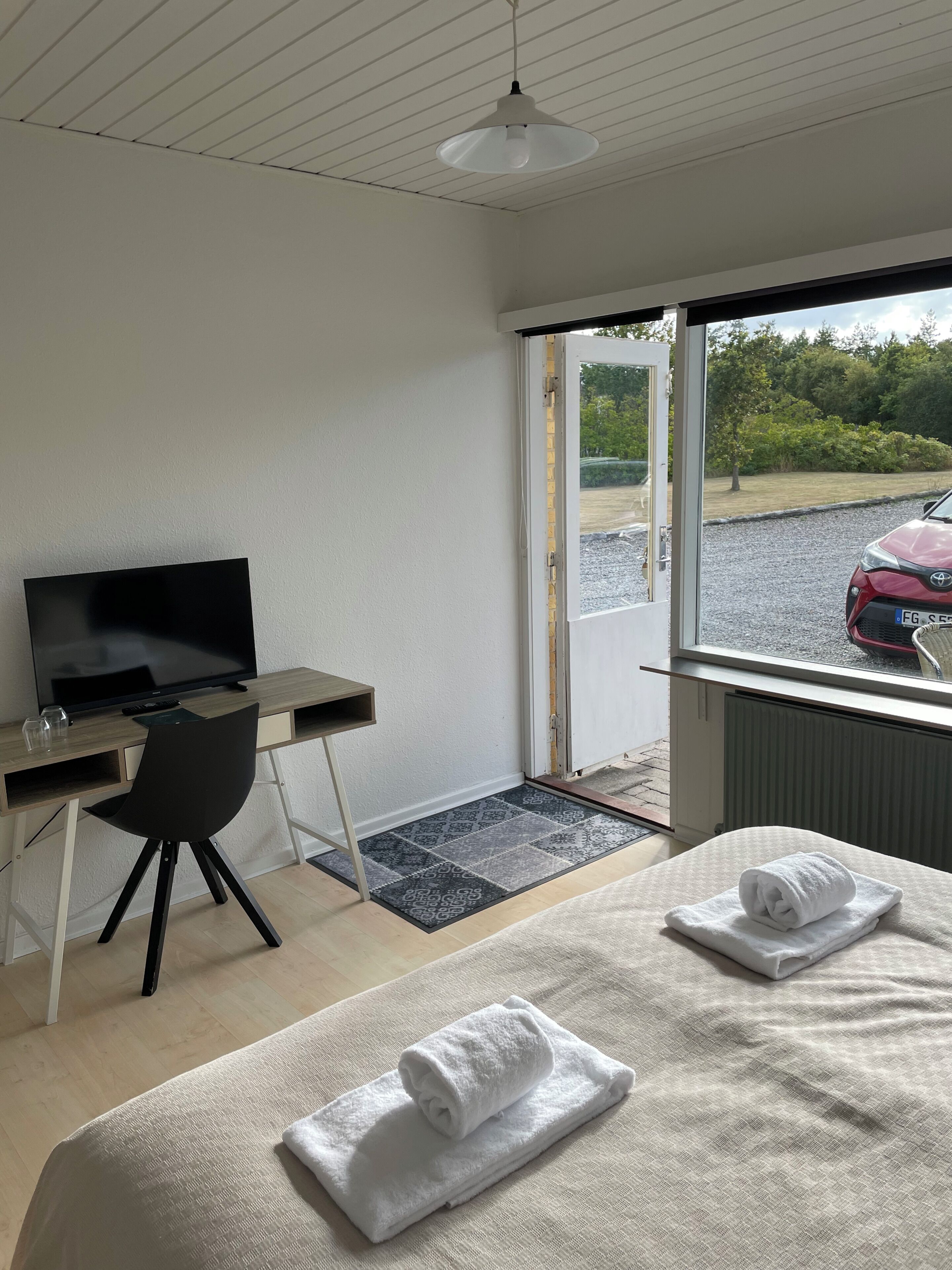 Habitación de hotel con cama, escritorio con televisor, una silla y ventana con vista a un coche y follaje exterior.