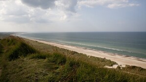 Un paisaje costero con una playa arenosa, dunas cubiertas de hierba y vista al océano bajo un cielo nublado.