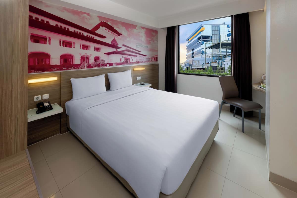 A hotel room with a large bed, a nightstand with a phone, a chair, and a view of a modern building through the window.