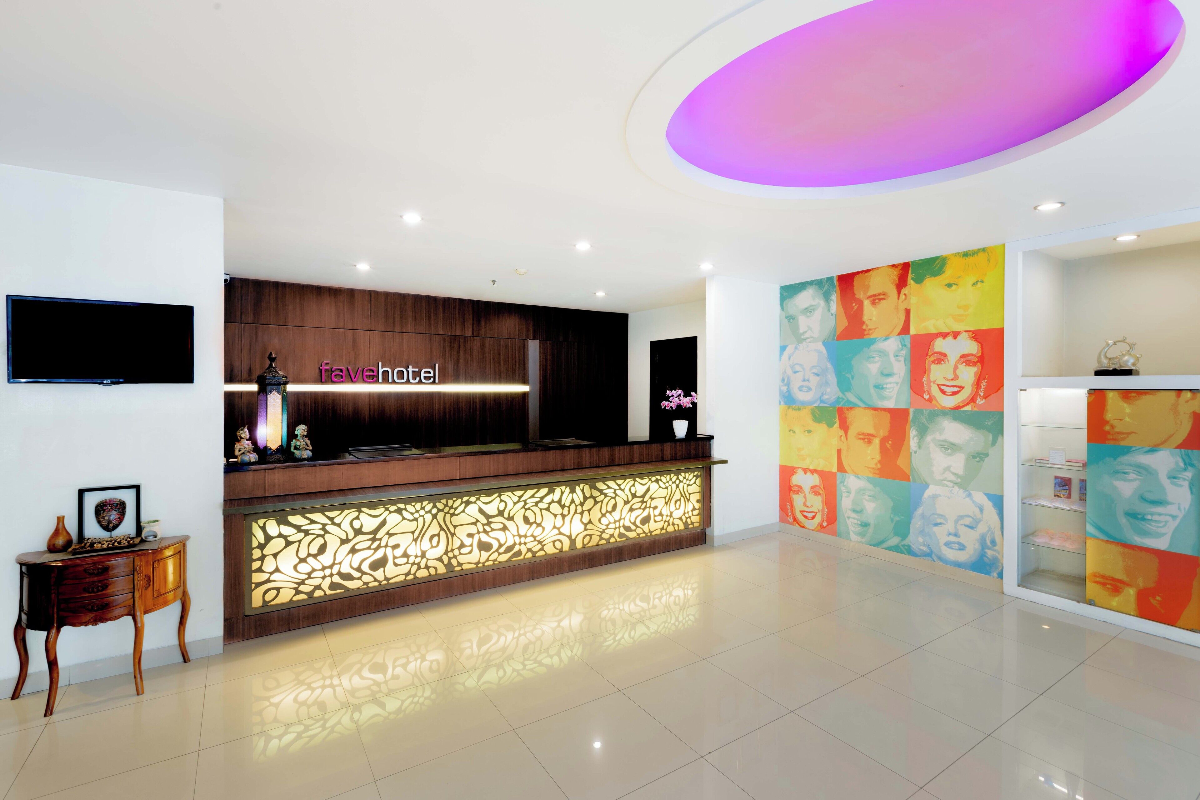 Zona de recepción de hotel con diseño moderno, que incluye un mostrador de madera, un televisor de pantalla plana y una pared colorida con arte pop.