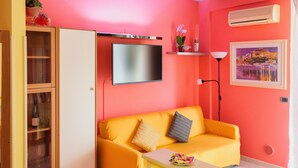 A room with a yellow sofa, a flat-screen TV, a small table with a bowl of fruit, a cabinet, and a wall-mounted air conditioner. - Patty's Aparthotel Arezzo (Arezzo)