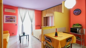 A brightly colored room with a bed, a dining table with a bottle and glasses, a clock, and a microwave on a shelf. - Patty's Aparthotel Arezzo (Arezzo)
