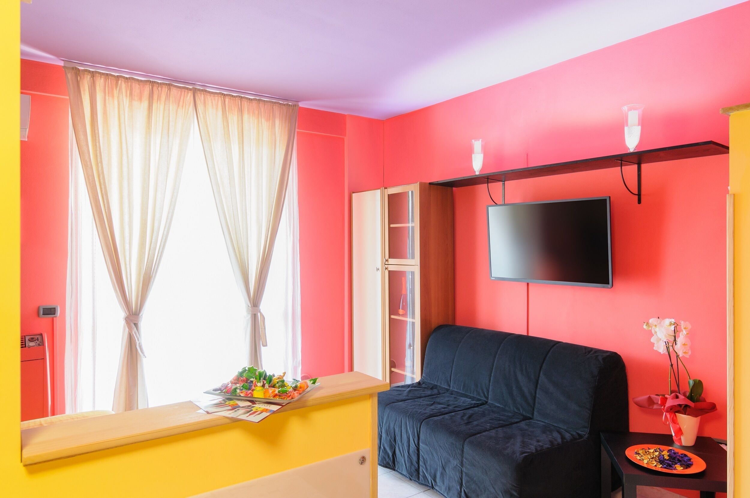 A room with a red wall, a black sofa, a TV mounted on the wall, and a window with curtains.
