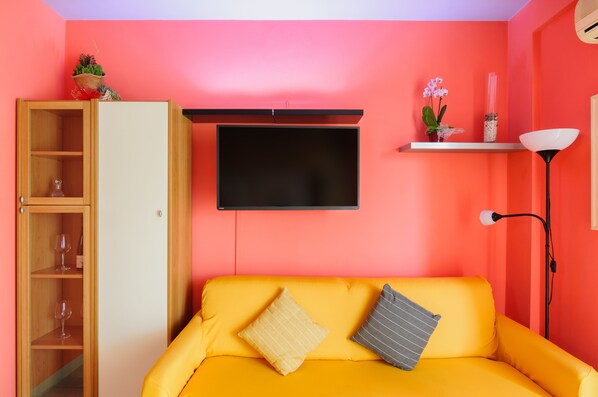 A room with a yellow sofa, a wooden cabinet, a TV mounted on the wall, and a small shelf with decorative items. - Patty's Aparthotel Arezzo (Arezzo)