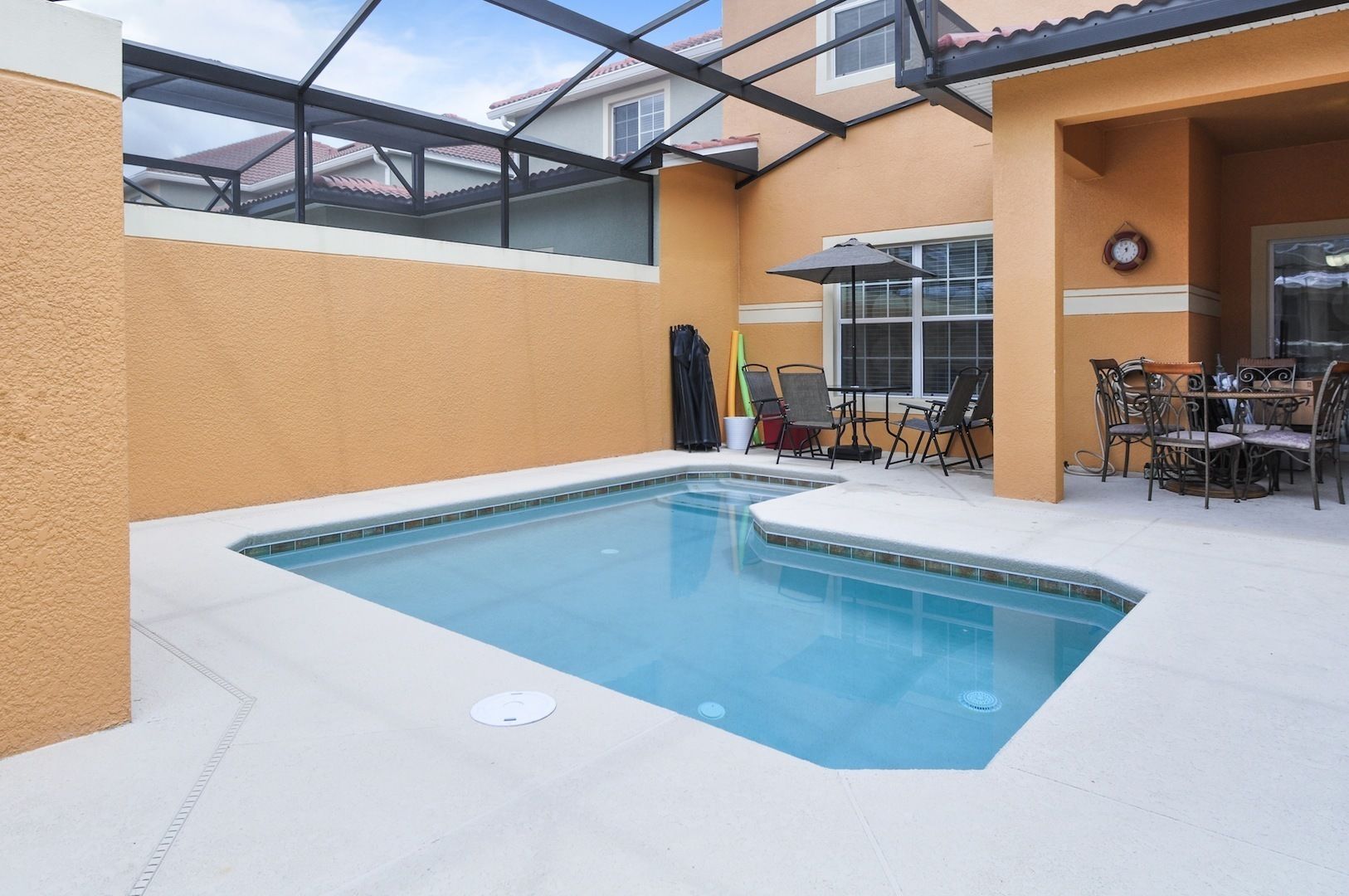 Una piscina con techo de cristal, rodeada por un muro color beige y un área de patio con mobiliario exterior.