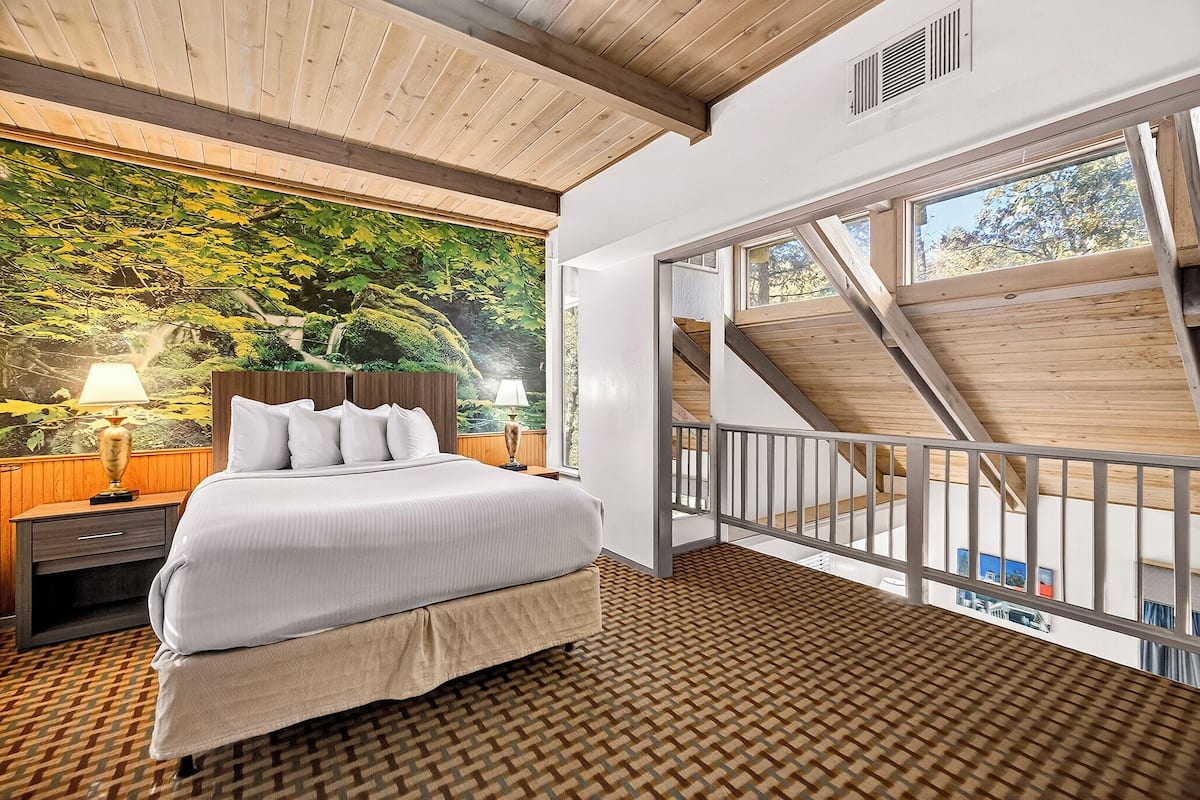 A bedroom with a bed, nightstand, and a mural of a forest. There is a balcony with a wooden railing and a view of the outside.
