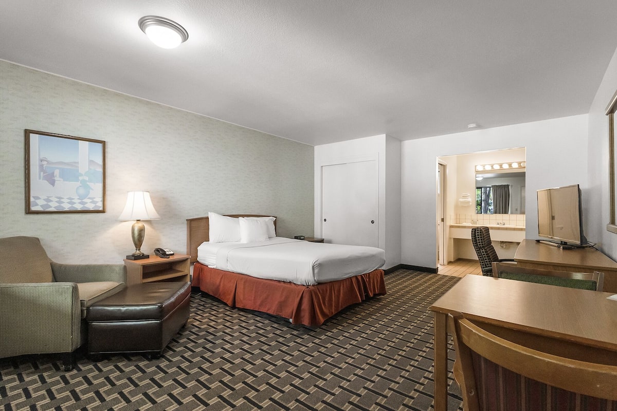 A hotel room with a bed, a desk, a chair, a lamp, a telephone, and a painting on the wall.