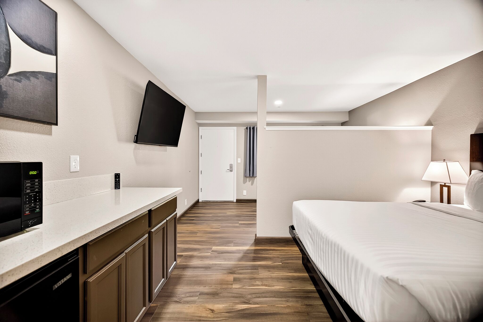 Une chambre d’hôtel moderne dotée d’un téléviseur à écran plat fixé au mur, d’un lit avec une literie blanche, d’une table de chevet avec une lampe et d’un plancher en bois.