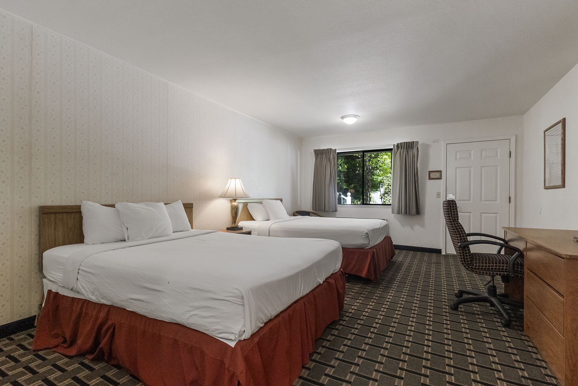 A hotel room with two beds, a desk, a chair, a lamp, and a window with curtains.