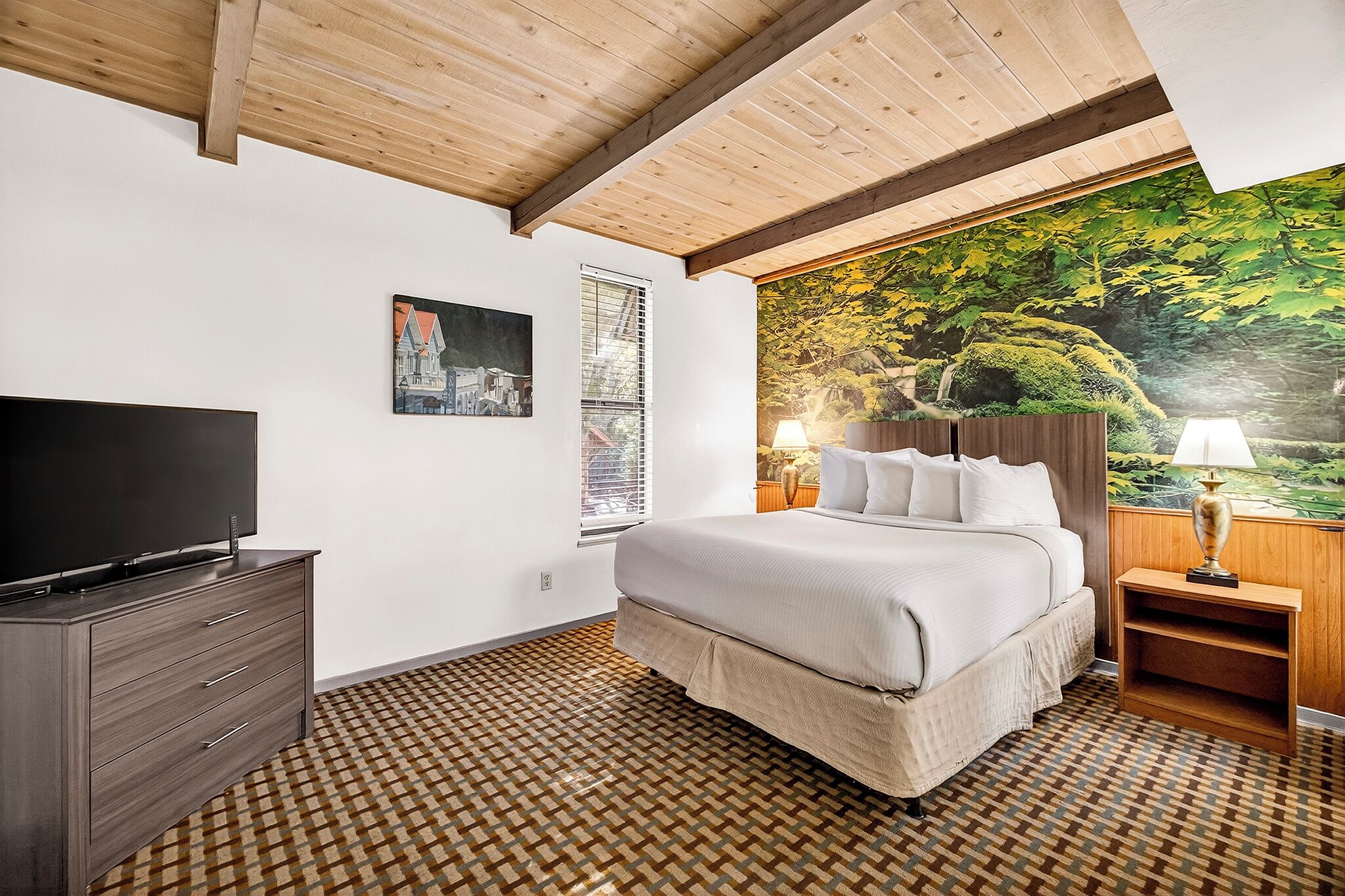 A bedroom with a large bed, a TV on a dresser, a mural of a forest, and a patterned floor.