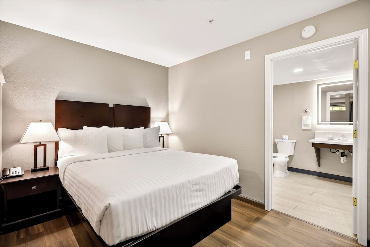 A hotel room with a bed, bedside tables, a lamp, a nightstand, a bathroom with a toilet, sink, mirror, and a door leading to another room.