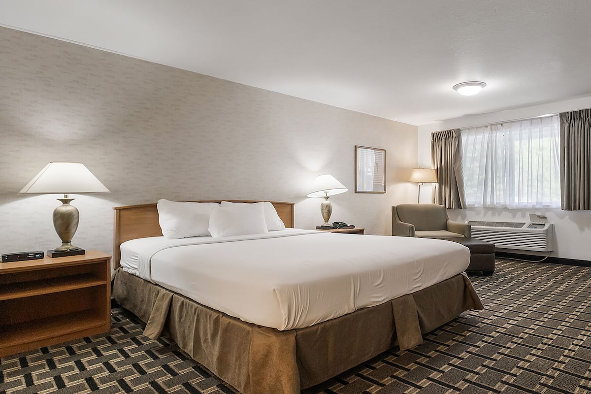 A hotel room with a large bed, two bedside tables with lamps, a chair, an air conditioning unit, and a door.