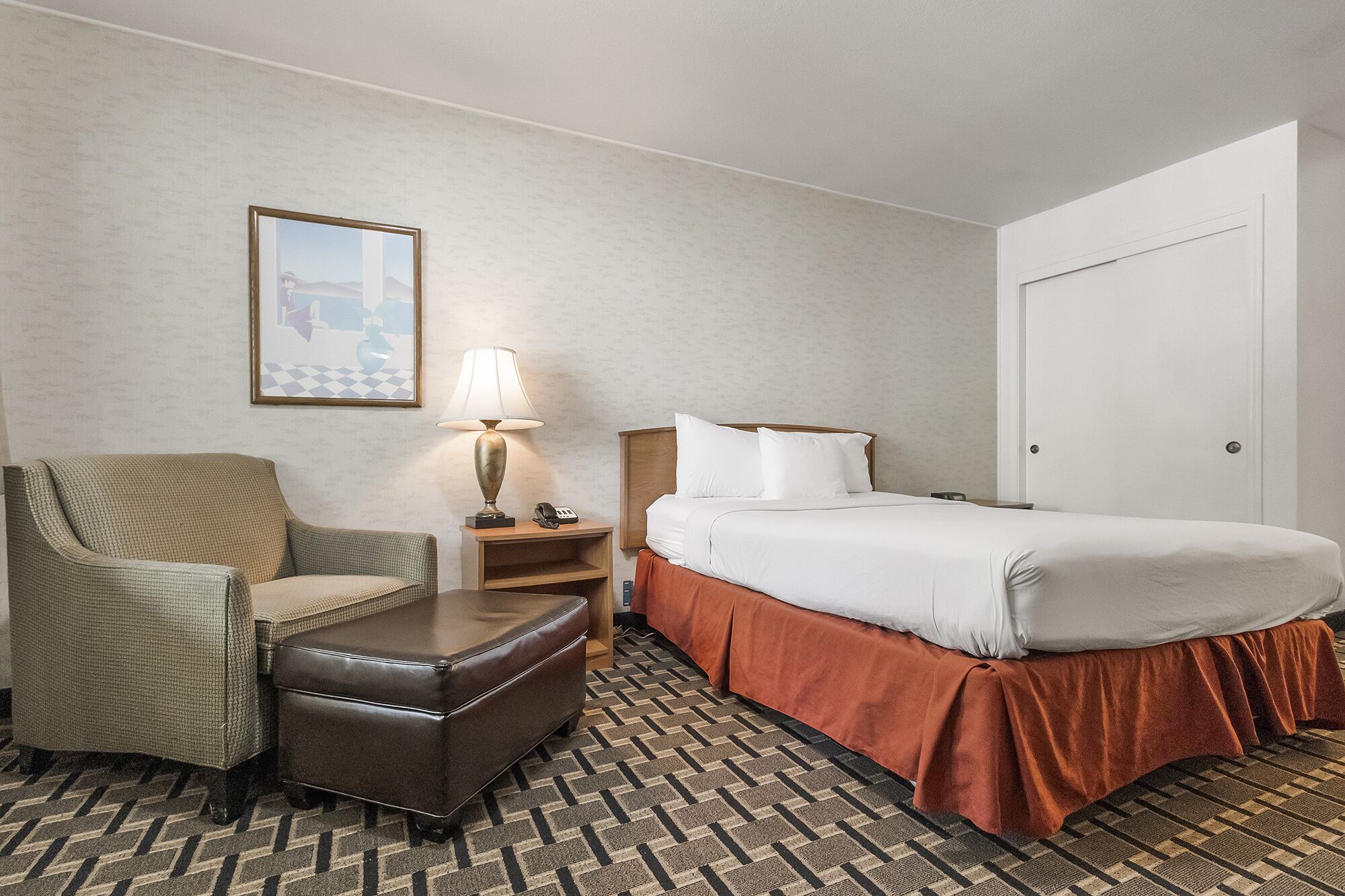 A hotel room with a bed, a chair, a nightstand with a lamp, and a framed picture on the wall.