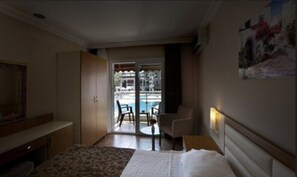 A hotel room with a bed, a desk, a chair, a small table, and a view of a pool area.