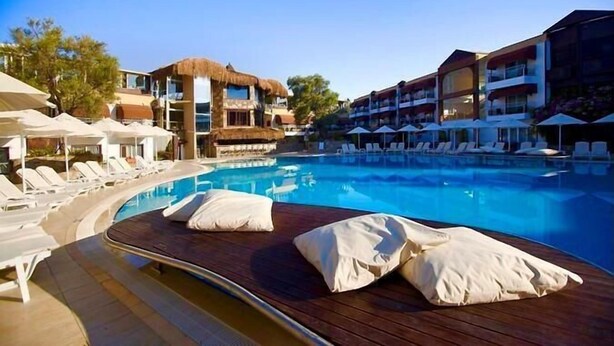 Un espace piscine avec des chaises longues et des parasols, un bâtiment à toit de chaume et des structures hôtelières modernes.