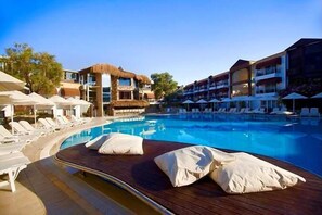 A pool area with lounge chairs and umbrellas, a thatched-roof building, and modern hotel structures.