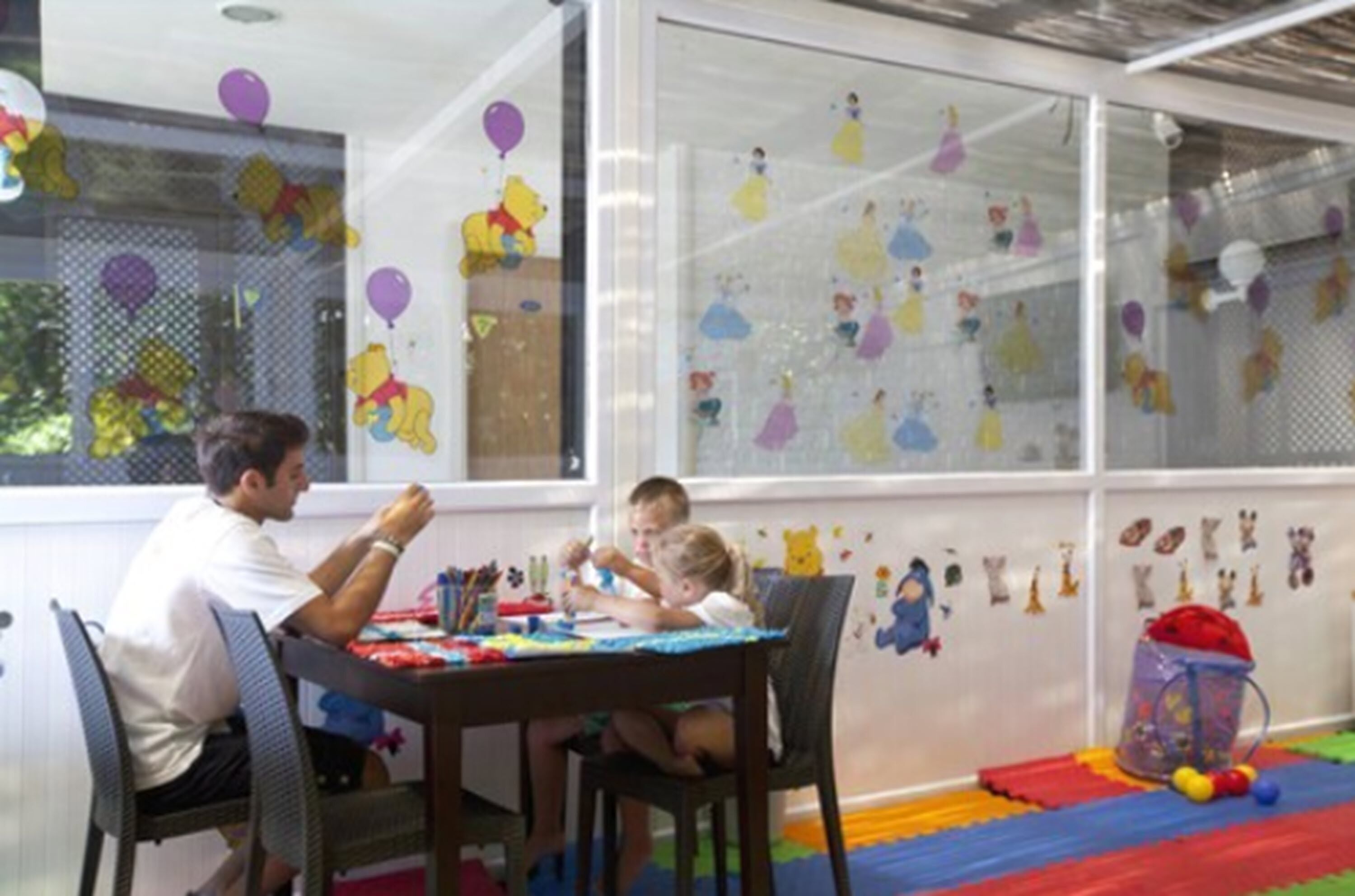 A room with a large window decorated with balloons and wall art, a table with children's toys, and a colorful play mat on the floor.