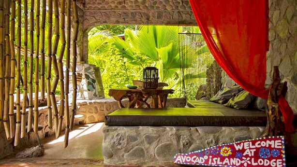 A tropical room with a stone bench, a small table, and a birdcage. A welcome sign is visible.