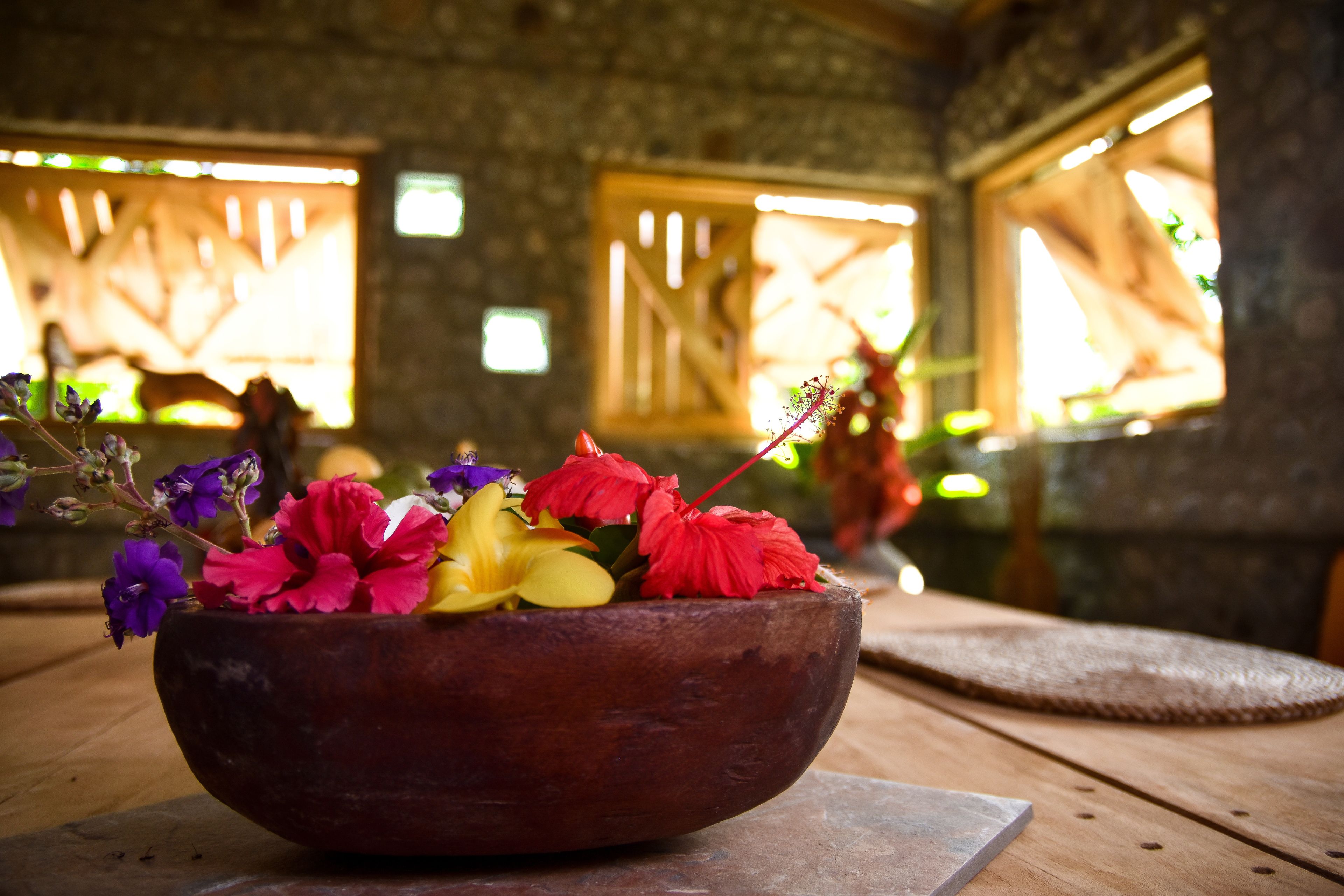 A wooden bowl filled with colorful flowers on a wooden table in a room with stone walls and windows.
