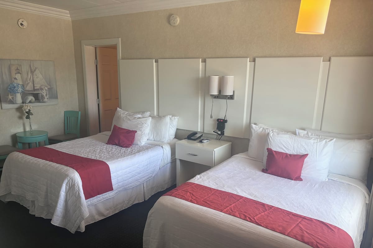 A hotel room with two beds, each with white bedding and red accents, a small round table with a chair, a nightstand with a phone and lamp, and a painting on the wall.