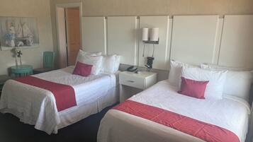 Ein Hotelzimmer mit zwei Betten, jeweils mit weißer Bettwäsche und roten Akzenten, einem kleinen runden Tisch mit Stuhl, einem Nachttisch mit Telefon und Lampe sowie einem Bild an der Wand.