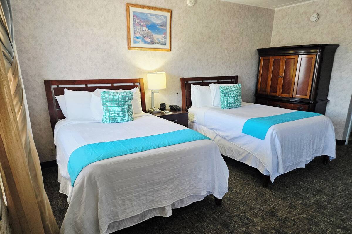 A hotel room with two beds, each with white bedding and teal accents, a wooden headboard, a nightstand with a lamp, and a painting on the wall.