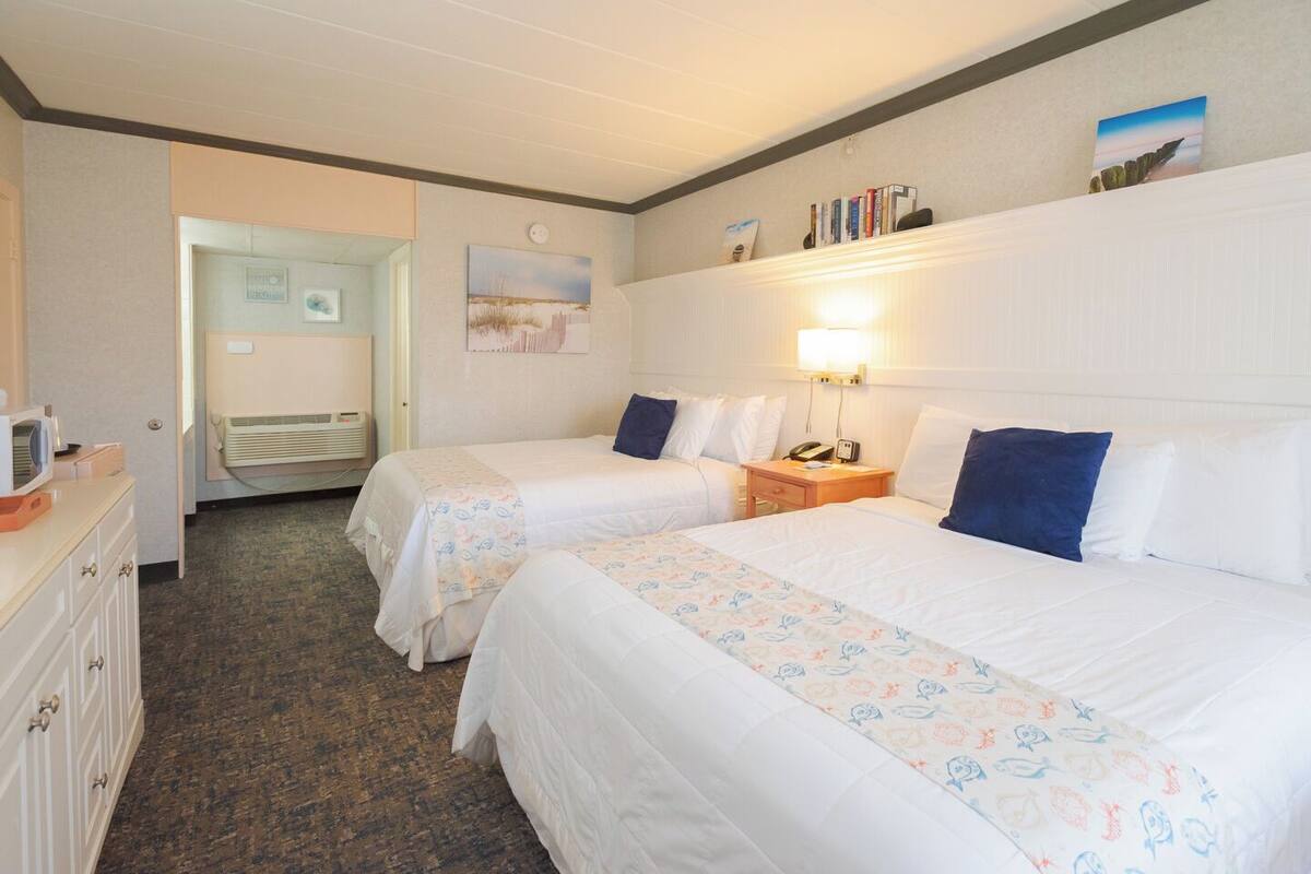 A hotel room with two beds, a desk, a chair, a lamp, and a bookshelf.