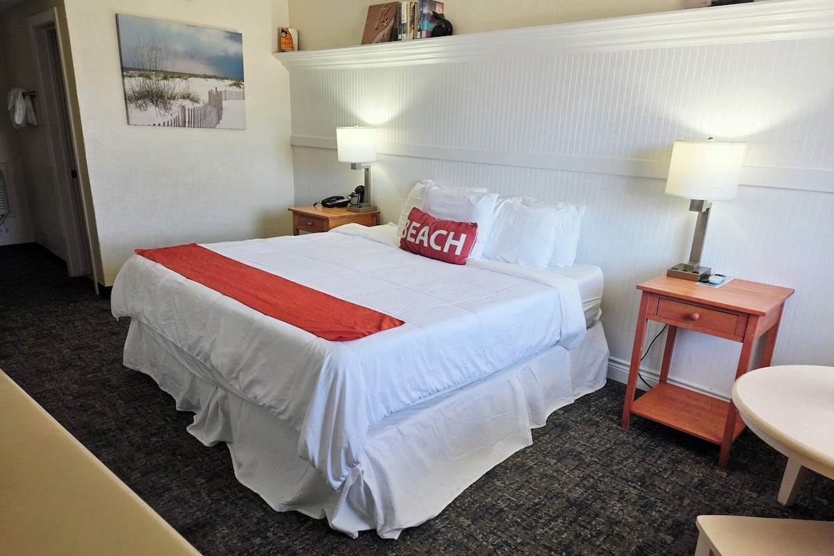 A hotel room with a large bed, two bedside tables, a chair, a painting on the wall, and a lamp on each bedside table.