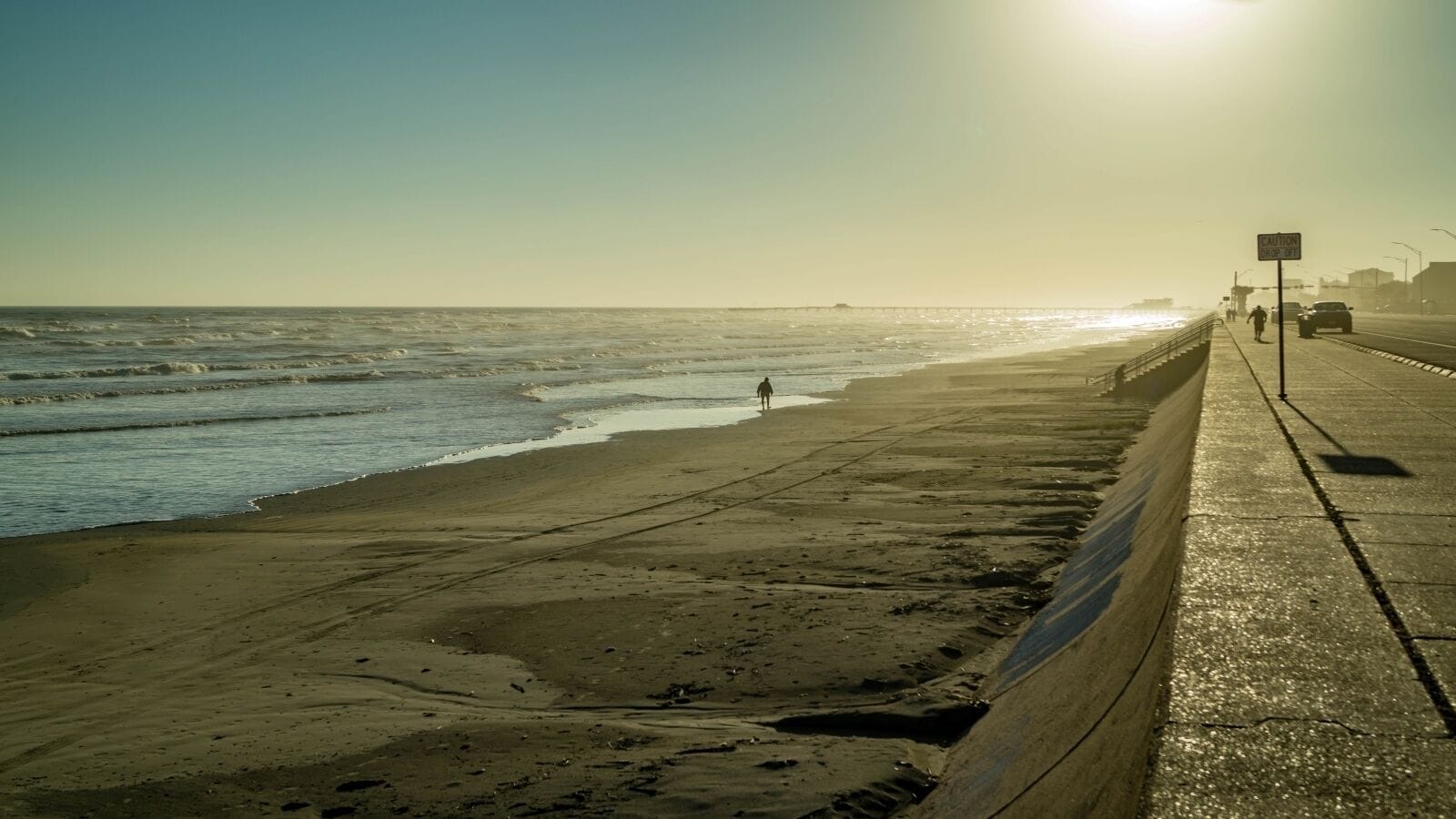 A beach with a concrete walkway, a person walking, and a distant pier.