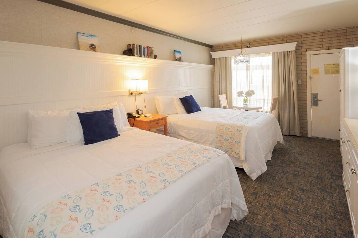 A hotel room with two beds, a bedside table with a lamp, a small table with a vase, and a door with a sign.