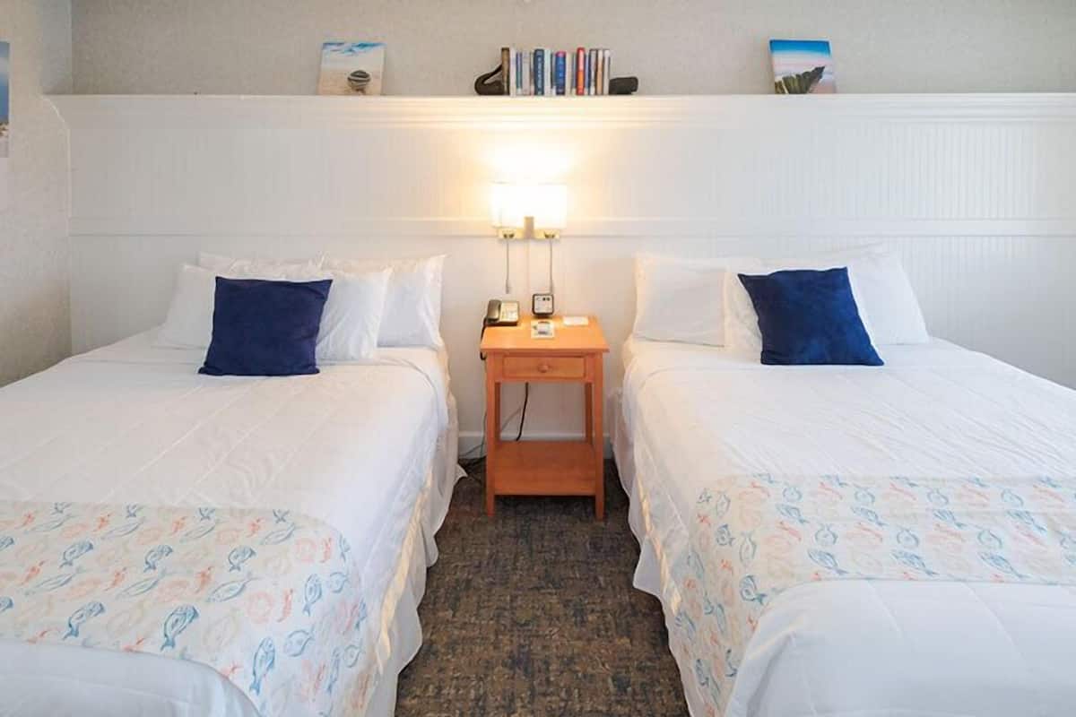A hotel room with two single beds, a small wooden nightstand with a lamp, and a shelf above the bed with books and a small decorative item.