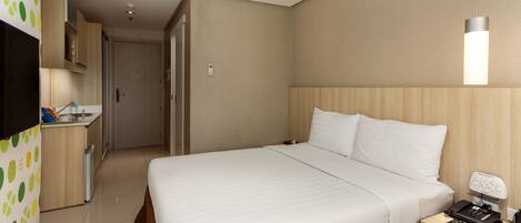 Quarto de hotel com uma cama grande, mesa de cabeceira com telefone, escrivaninha com luminária, pequeno espaço de cozinha e televisão.