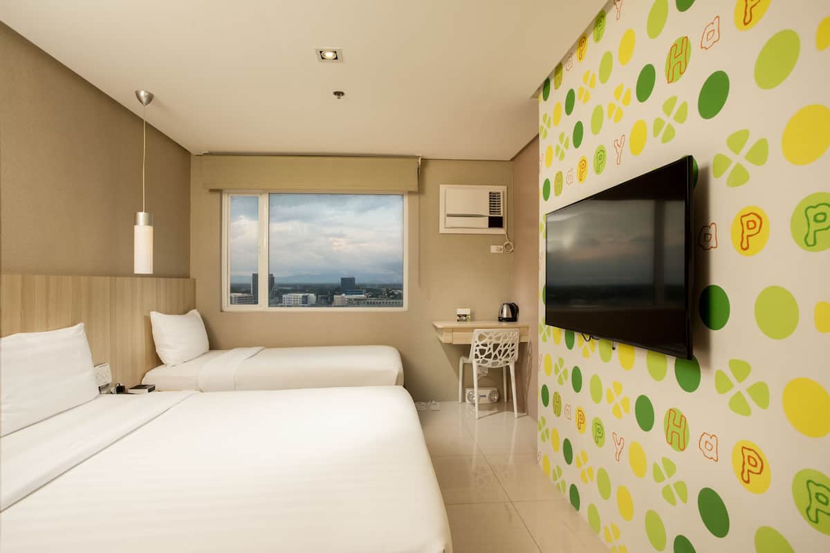 Ein Hotelzimmer mit zwei Betten, einem Schreibtisch, einem Fernseher und einem Fenster mit Blick auf die Stadt.